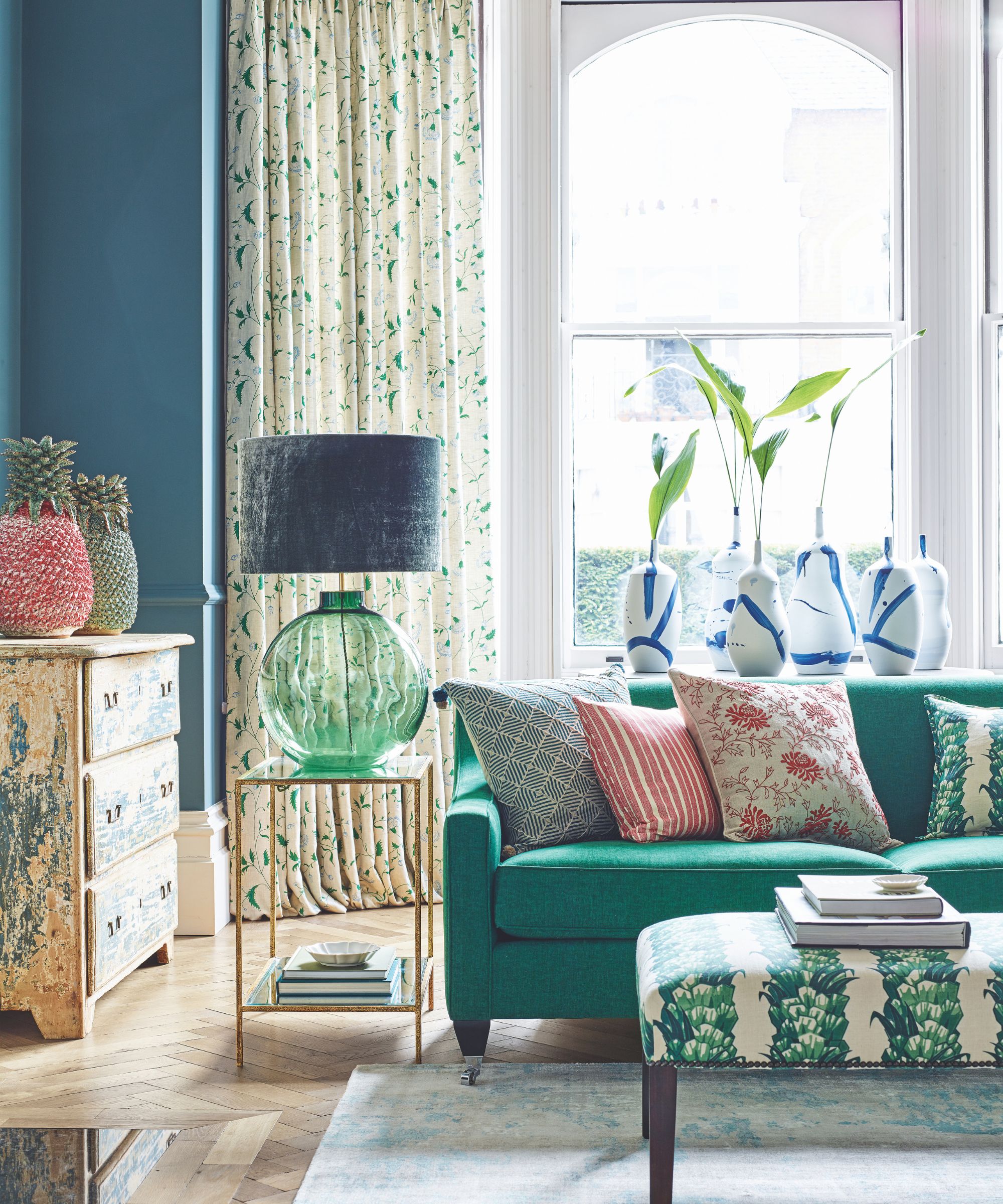 A bright and colorful living room with layered patterns, with a green sofa, a patterned rectangular foot rest, colorful cushions, a green glass lamp with a blue velvet shade, a patterned chest of drawers and large bay windows behind, with patterned curtains.