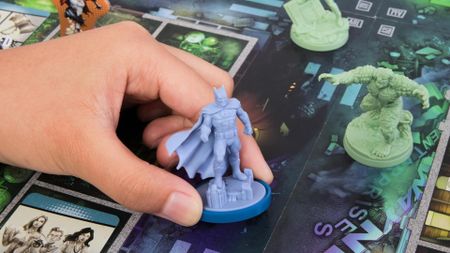 A hand holds a Batman miniature over a board with a Killer Croc model visible