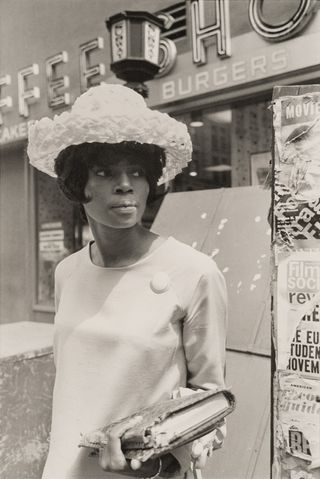 A black and white photo of a woman with a short bob wearing a light coloured hat and outfit standing outside a shop. She is holding a book in one hand