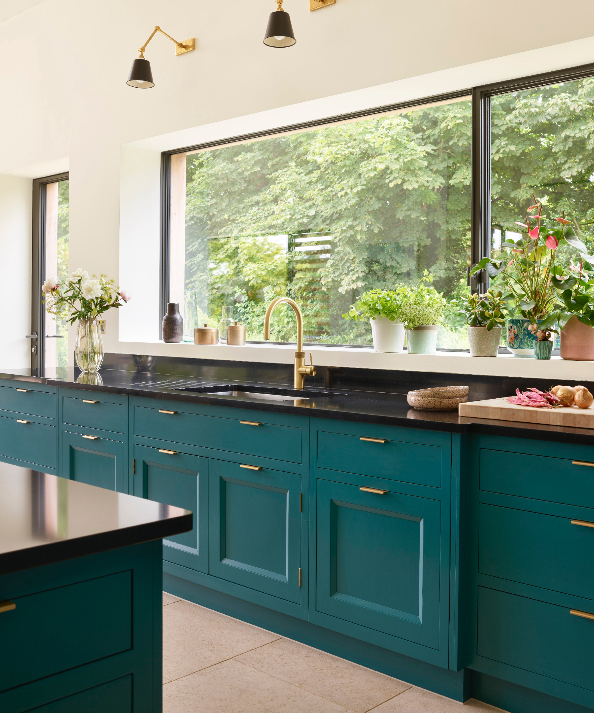 teal kitchen with black counterops and island top, black and brass wall lights, brass faucet, crittall windows, plants on windowsill