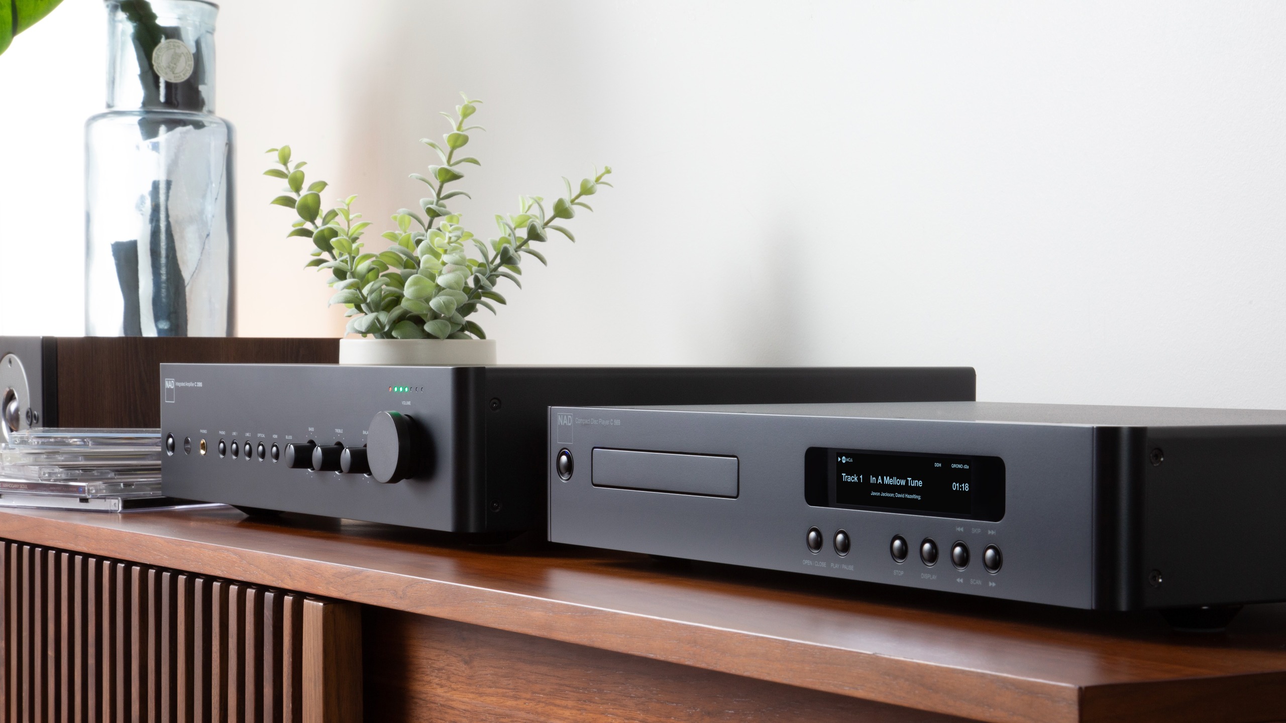 The NAD C 359 streaming amplifier and C 569 CD player in black on a wooden cabinet, seen from a 3/4 angle.