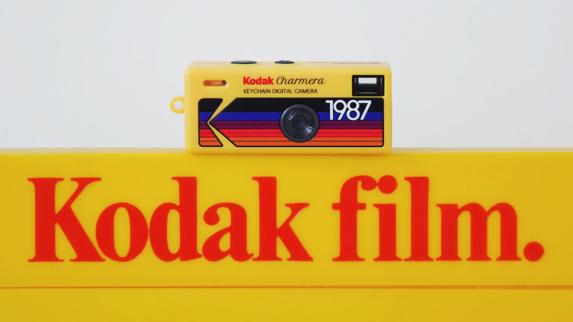 A vintage Kodak Charmera digital camera from 1987 sits atop a bright yellow Kodak film sign, showcasing retro photography nostalgia