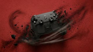 A black gaming controller shatters dramatically against a red background, surrounded by swirling debris, conveying motion and intensity.