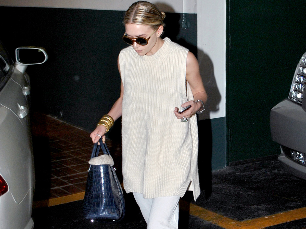 Ashley Olsen walking in a car garage in Los Angeles with a slicked back bun wearing round sunglasses, a ribbed sleeveless tunic sweater, Sidney Garber gold bracelets, a blue croc The Row bag, and white linen pants.