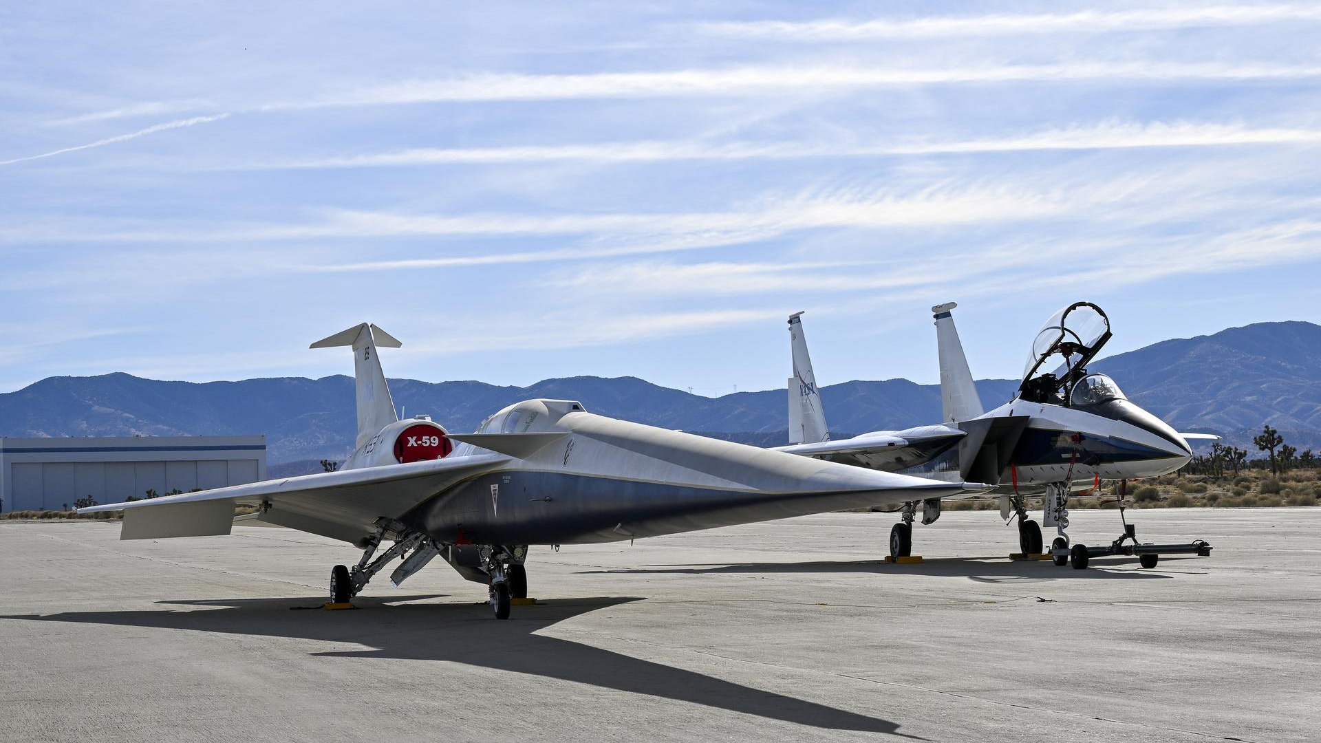 A tall gray plane with an open cockpit sits to the right of a sleeker, shorter plane with a red flag labeled X-59 over its cockpit. Both planes sit on a sunny tarmac