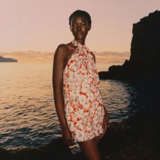 A model standing in front of a sunset over the ocean wearing a halter neck NET-A-PORTER red and white floral dress. 