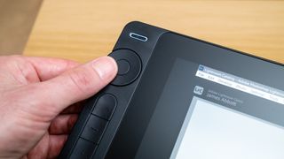 Thumb pressing a button on a Huion Kamvas 16 (Gen 3) drawing tablet on a wooden desk