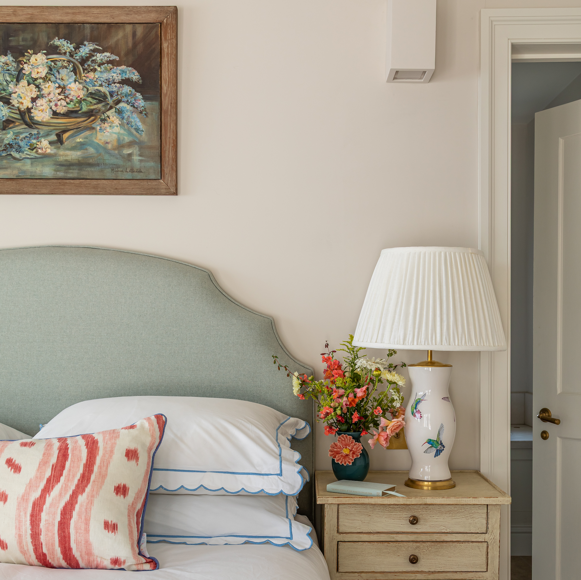 bedroom with pale green upholstered headboard beneath oil painting of flowers with lamp on bedside table