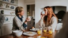 Two women sit at a table drinking from mugs, with eaten boiled eggs and glasses of orange juice on the table