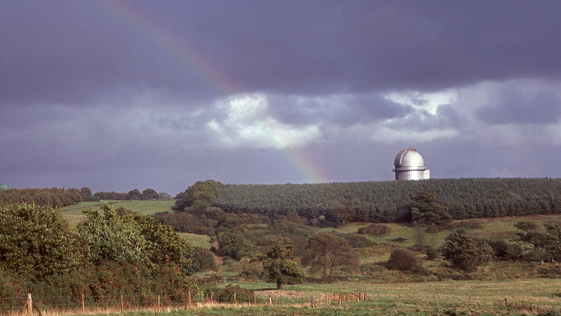 a rainbow stretches across the sky above a domed building in in a lush green countryside