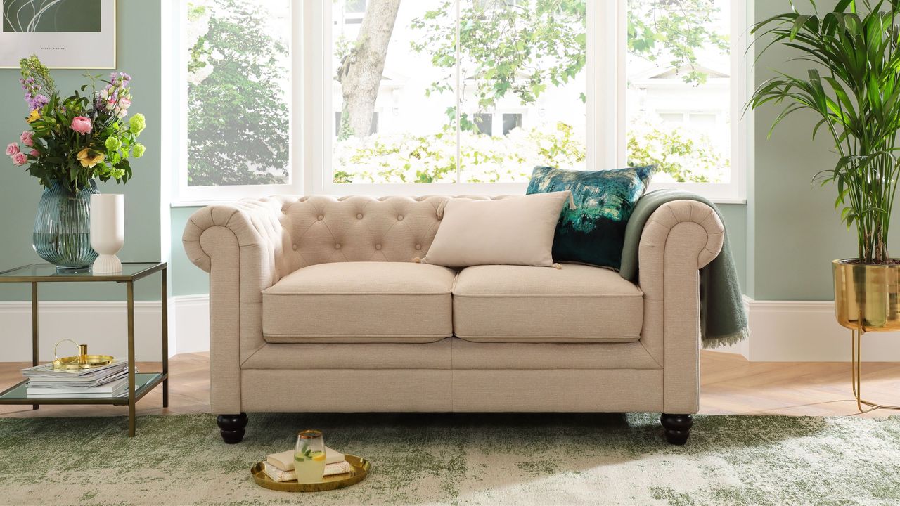 Things people with nice-smelling homes never do is useful to know. Here is a clean home - a living room with sage green walls, a large window reflecting trees and white buldings, a beige couch with throw pillows, a gold side table with flowers on, and a green and white gradient rug