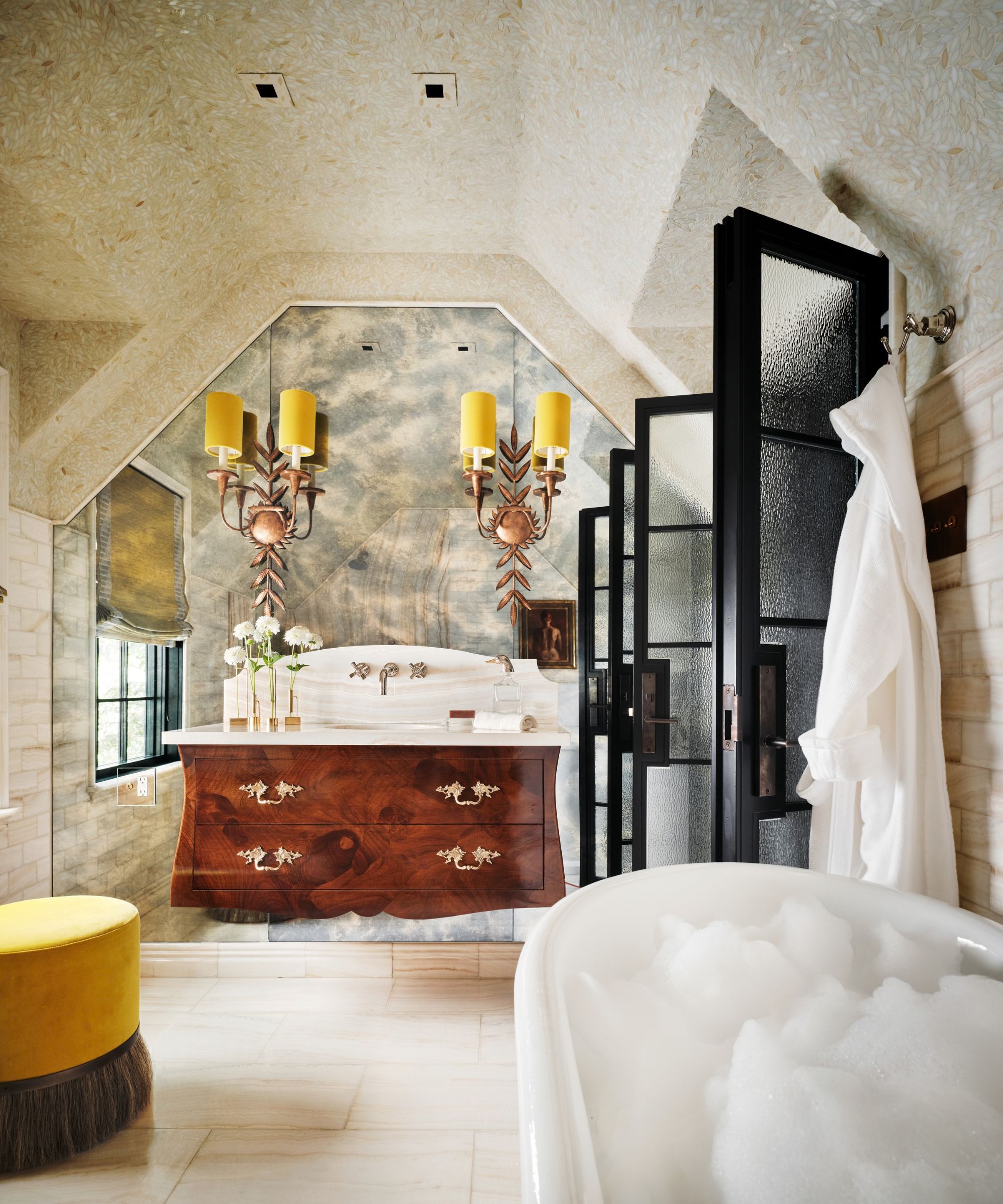 Grand bathroom with antique mahogany vanity unit, two antique candelabra sconces above with yellow shades, mosaic walls and ceiling, glass doors, a yellow ottoman, and a bathtub.