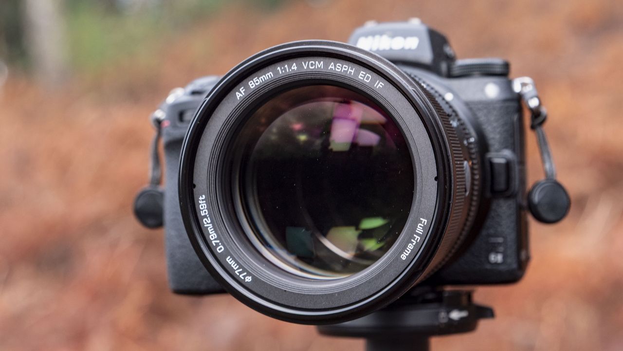 Close-up of the Viltrox AF 85mm F1.4 Pro lens attached to a Nikon camera, mounted to a tripod outdoors and with bronze-colored ferns in the background, emphasis on the lens aperture blades
