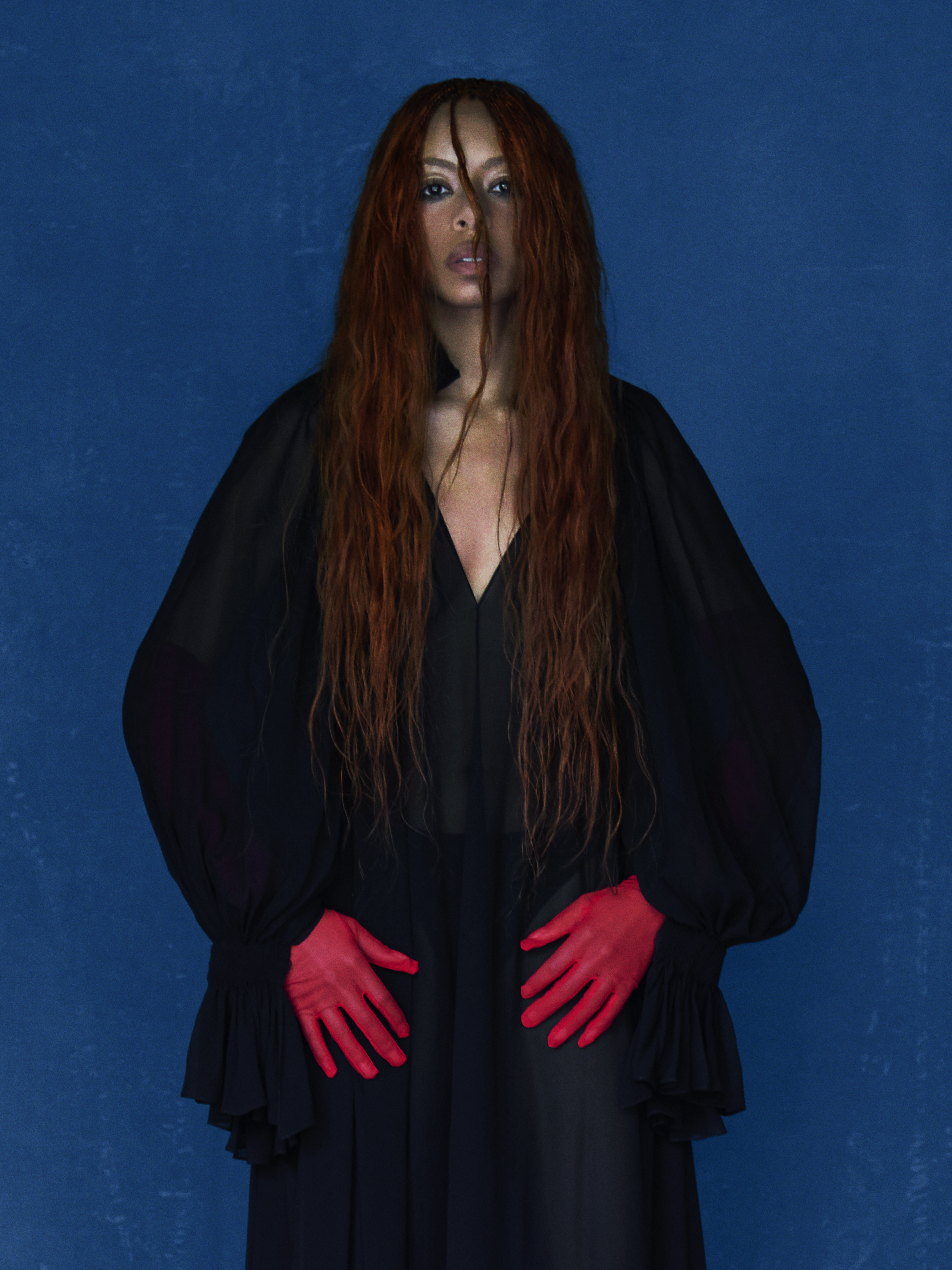 singer ravyn lenae poses against a blue backdrop wearing an all black outfit and red gloves in a promotional shot for her album blue island