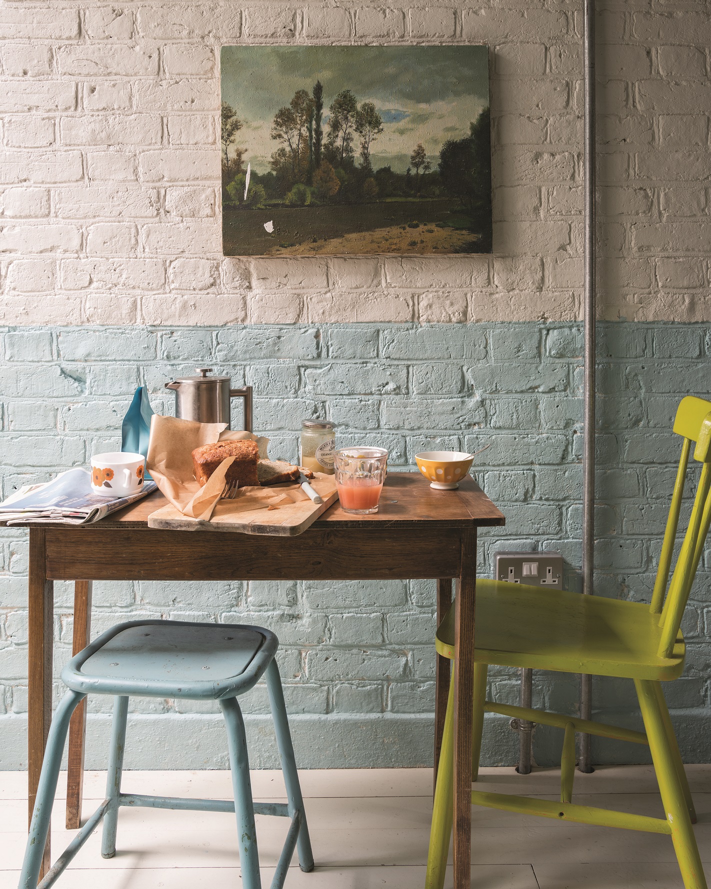 Exposed brick wall painted in blue and pink by Farrow and Ball with wall painting, plus a small dining table with green chair and blue stool