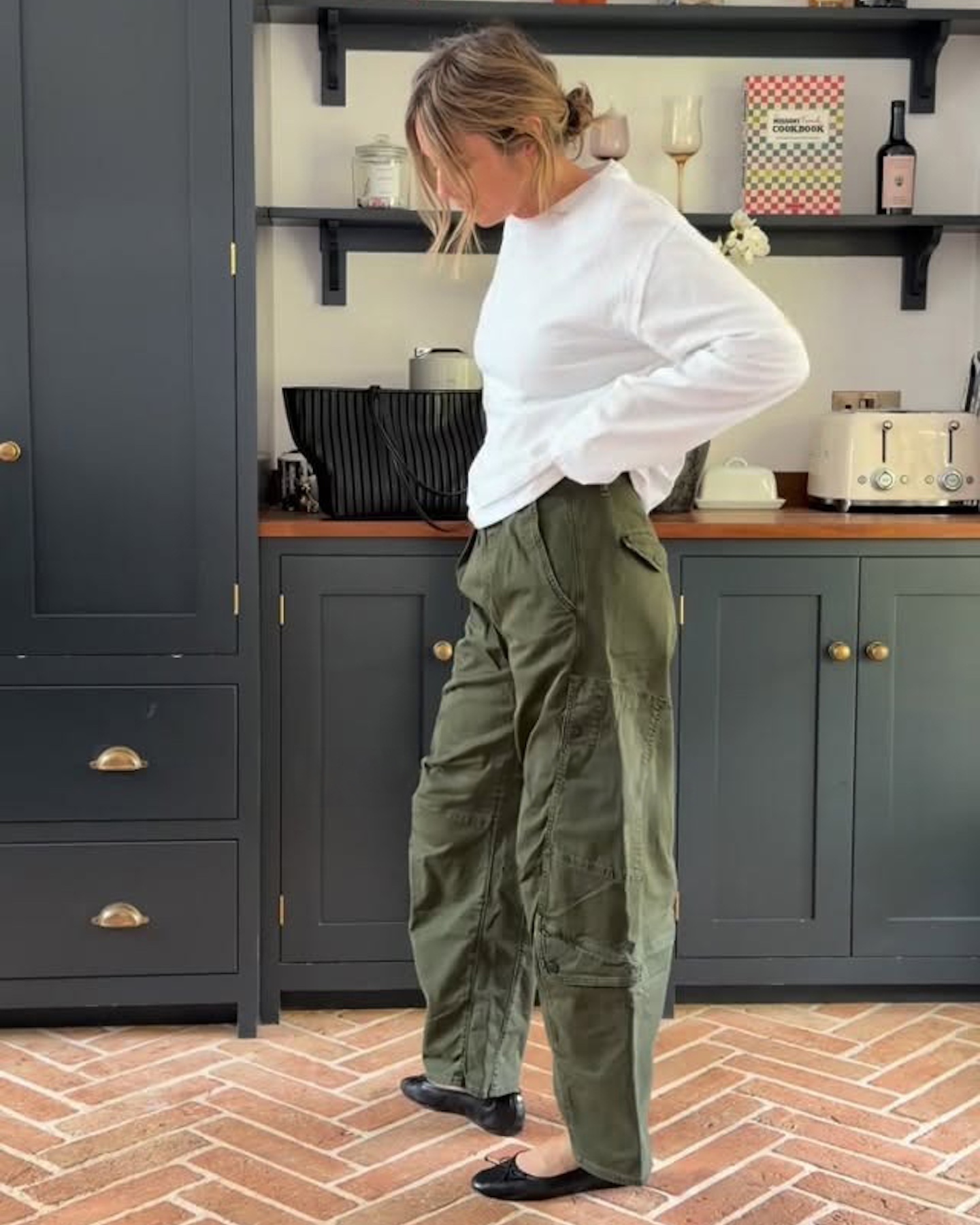 British style influencer Emma Rose Thatcher poses in her kitchen wearing an oversize long-sleeve white T-shirt, a woven black bag, green barrel pants, and black ballet flats