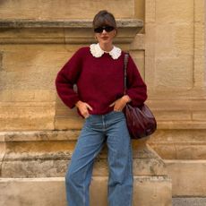 Influencer @juliesfi poses for a photo wearing a burgundy knit with blue jeans, brown loafers and a burgundy bag.