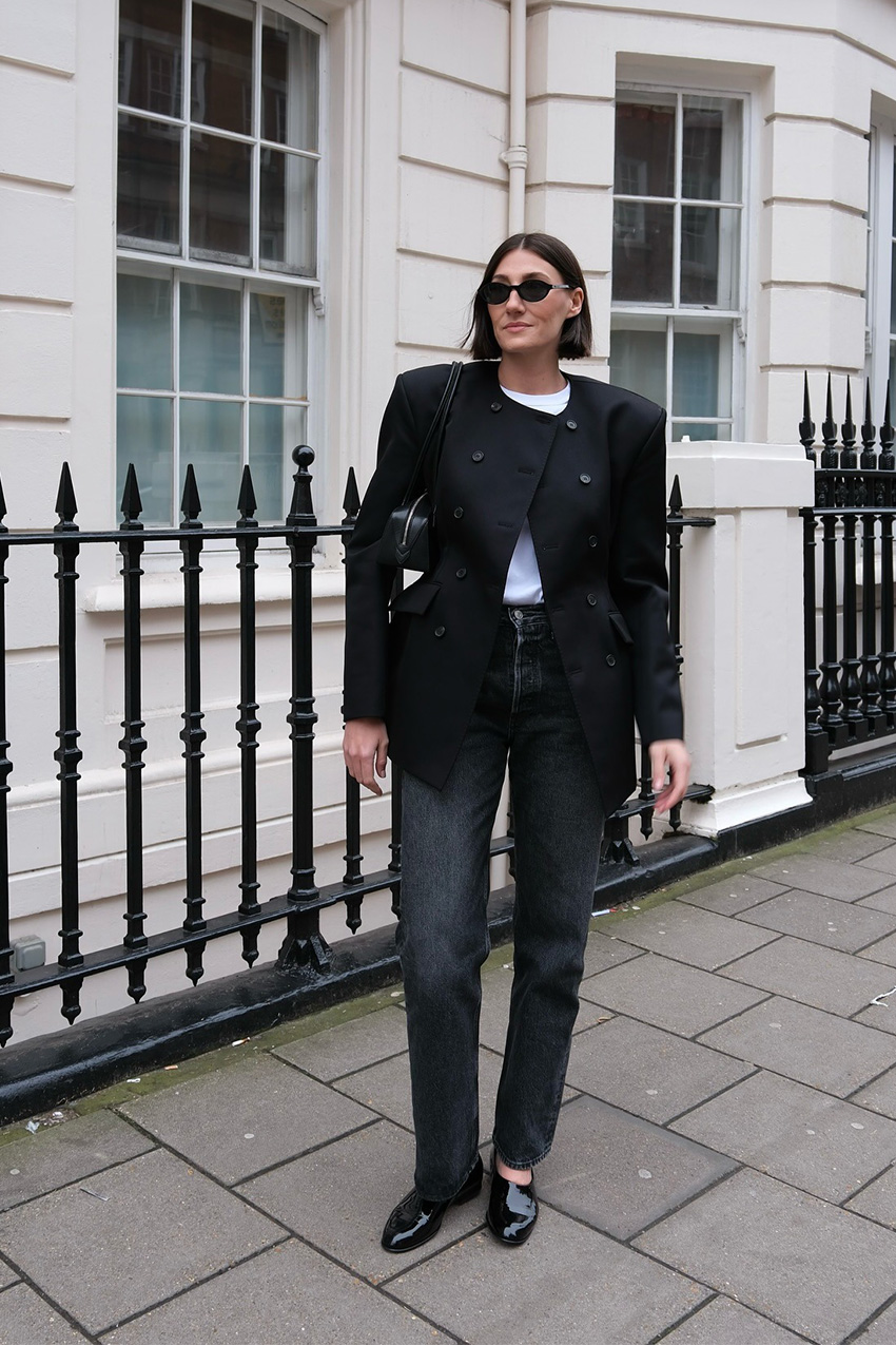 How to wear skinny jeans is shown in a photo of a woman standing outside on the sidewalk wearing black sunglasses, a black blazer over a white t-shirt styled with black slim straight-leg jeans, black flats, and a black shoulder bag
