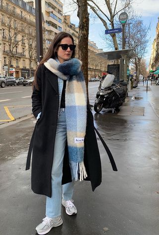 A French woman standing outside wearing a checkered cashmere scarf over a black coat, black sweater, baggy jeans, and chunky white sneakers.