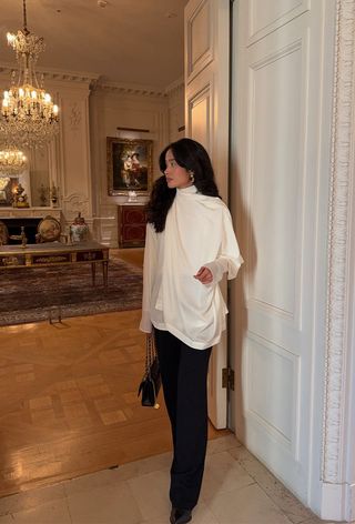 Hanukkah outfit idea is shown in a photo of a woman inside a beautiful home wearing gold earrings, a white draped blouse, black trousers, black pointed shoes, and a quilted black handbag