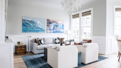 Coastal living room ideas. Coastal living room with two pieces of artwork on wall, light gray painted wall, large glass chandelier, blue rug over wooden floor, two cream armchairs facing a gray sofa with dark gray coffee table