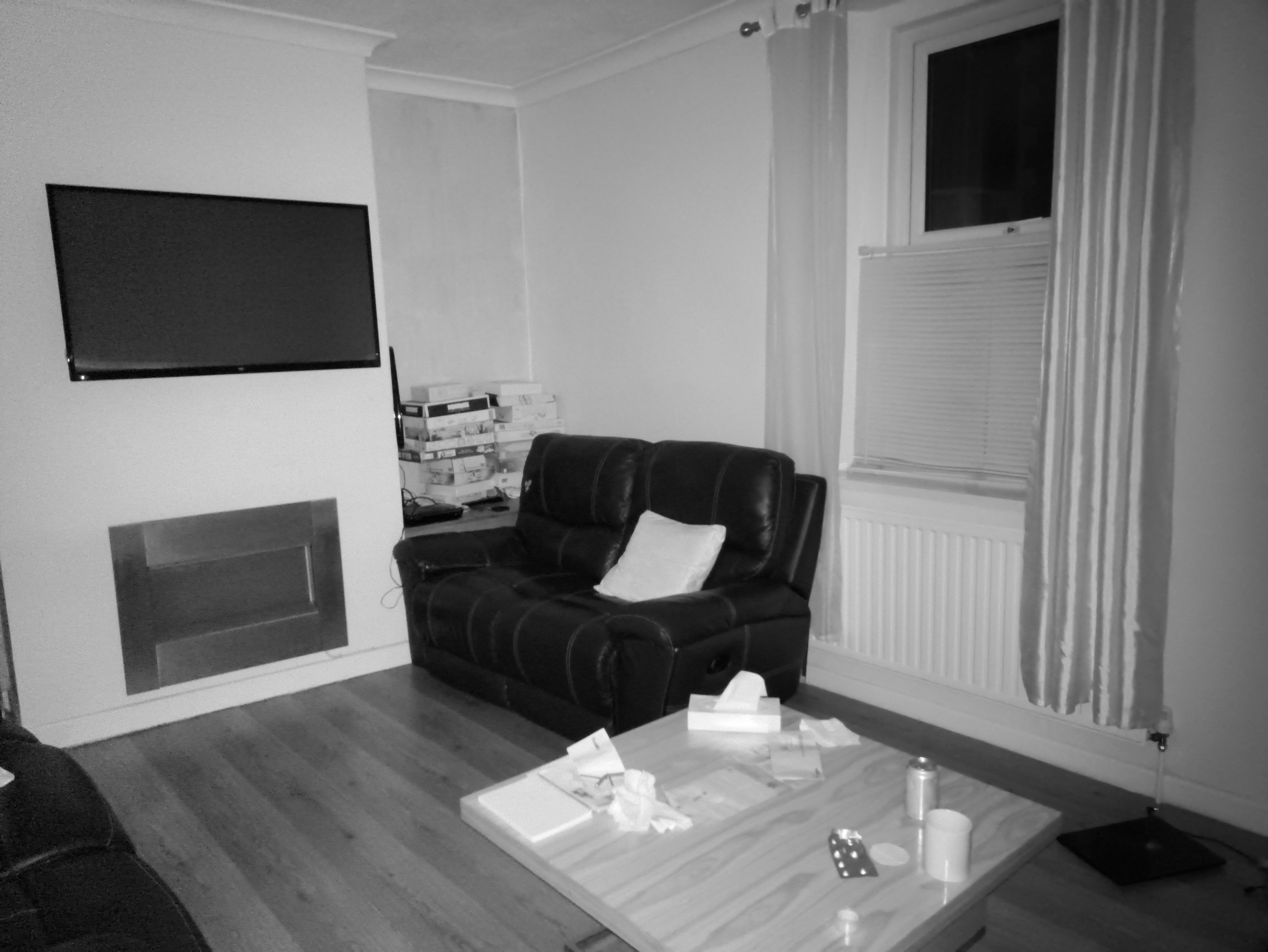 A living room corner featuring a wall-mounted television above a fireplace opening, a black leather reclining loveseat, and a square wooden coffee table with scattered items.