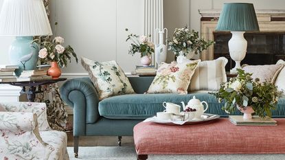 A living room with turquoise velvet sofa, pink upholstered footstool and floral white cushions