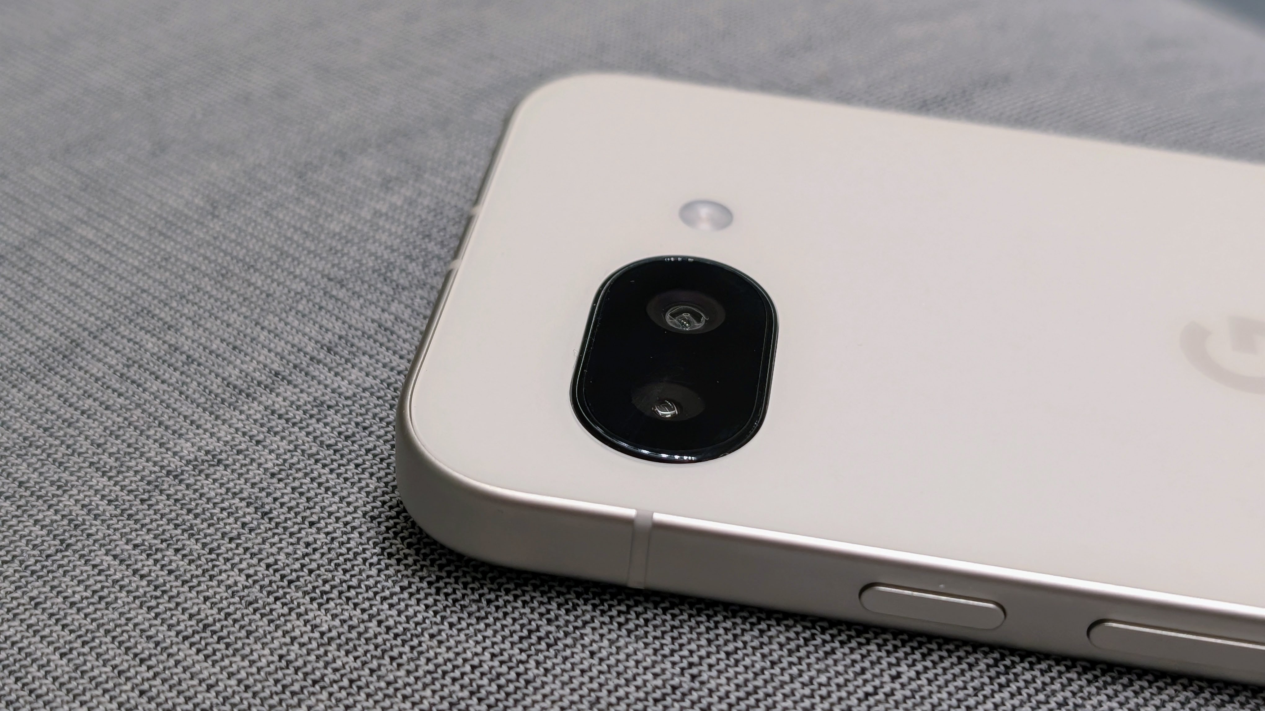 A close-up of the top of the Porcelain Google Pixel 9a, focused on the camera cutout with buttons and the Google logo visible.