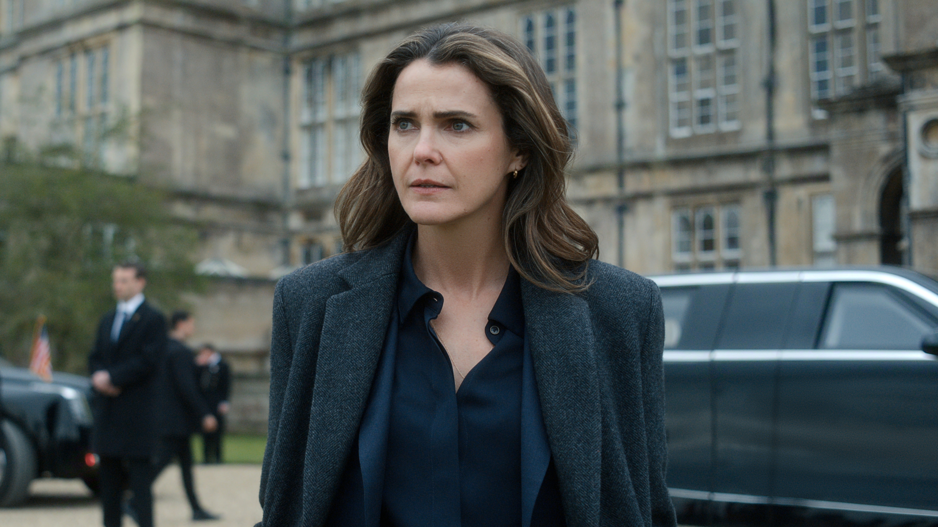 Keri Russell as Kate Wyler, standing in the driveway of a British castle, with suited men and black cars behind her, in 'The Diplomat' season 4.