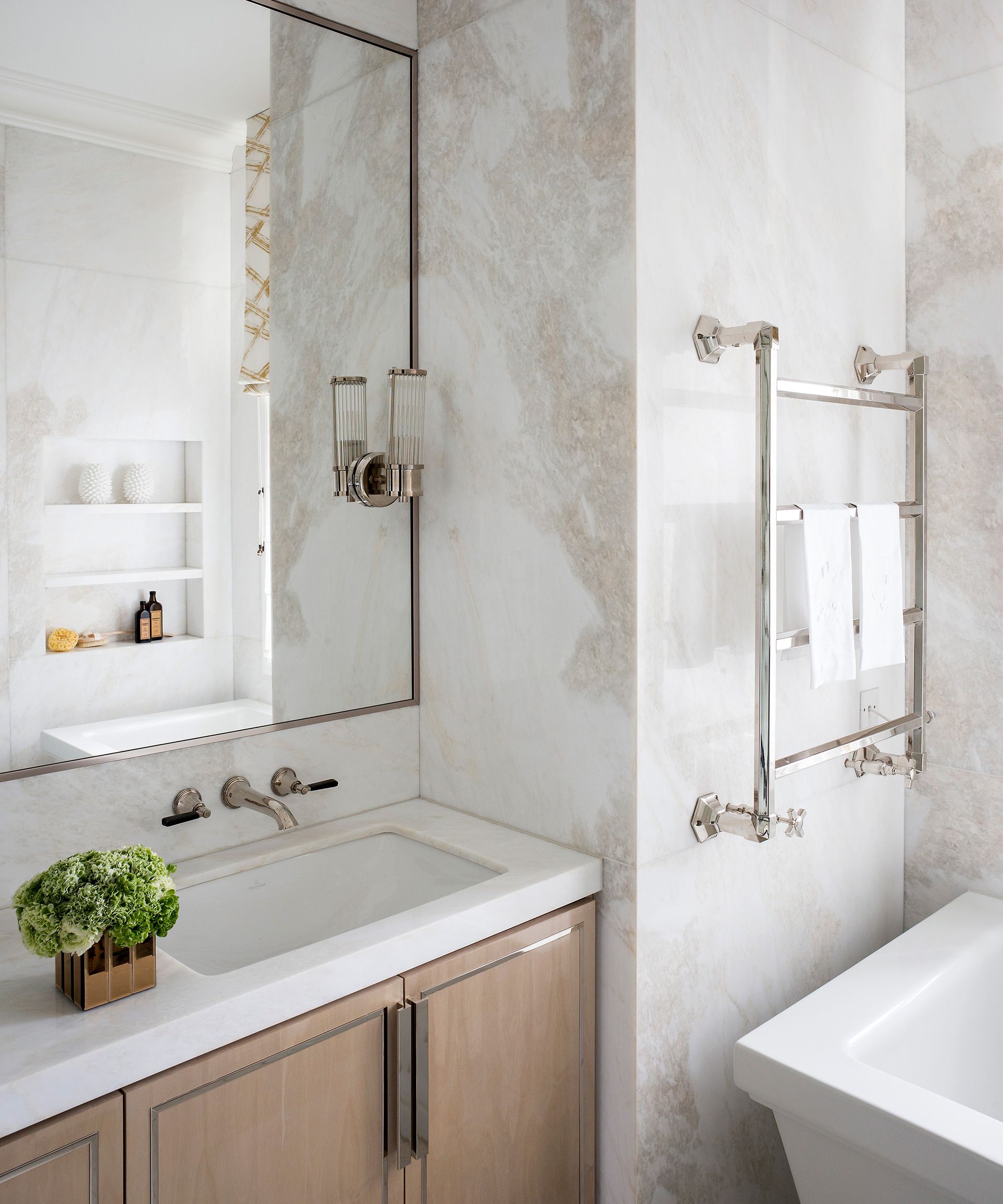 A small bathroom with a wall-mounted towel warmer radiator over a white square edged tub (corner visible). beside a wooden vanity with an inset white skin basin and a large wall mirror.