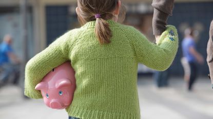 A little girl with a piggy bank under her arm holds her dad's hand.