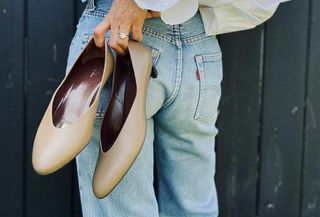 Woman holding up nude court shoes while wearing white button-down shirt and light-wash jeans.