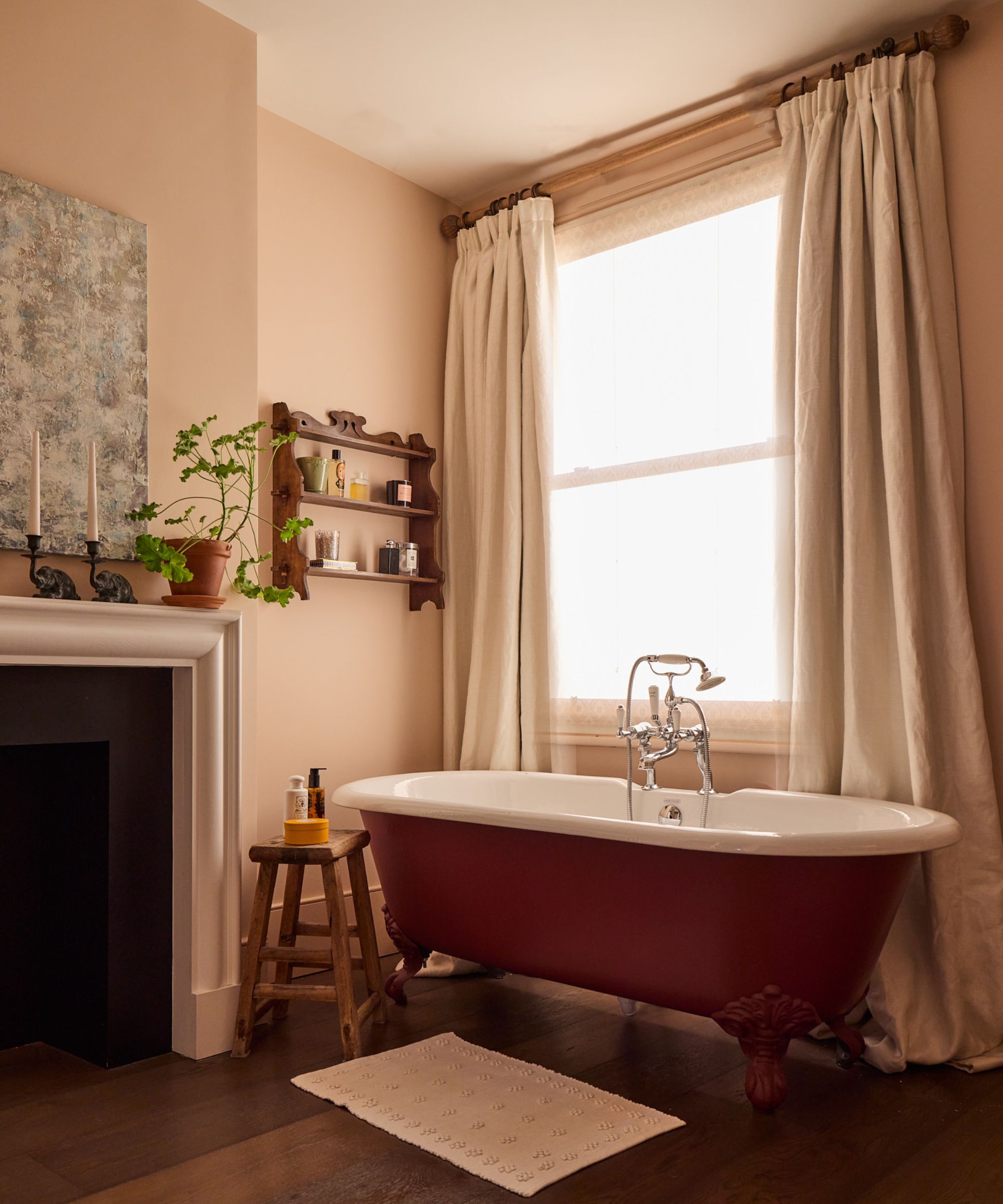 A traditional bathroom with plaster pink walls, a sash window with neutral curtains, a red freestanding bathtub and wooden flooring and a white fireplace with decorative items on the mantel.