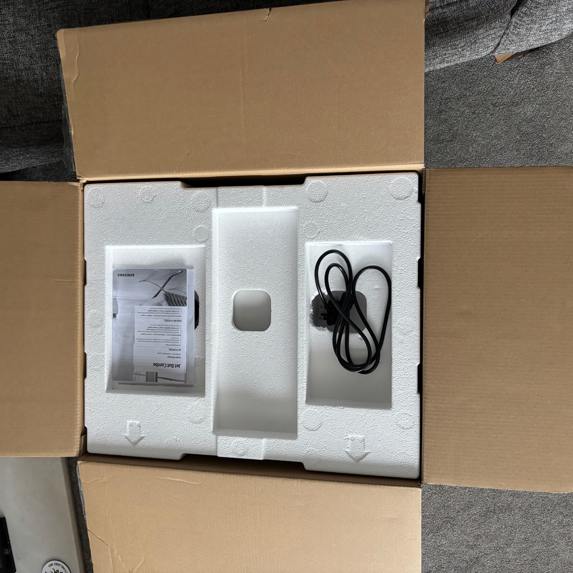 Opened cardboard box view featuring polystyrene layer with black cable and user manual 