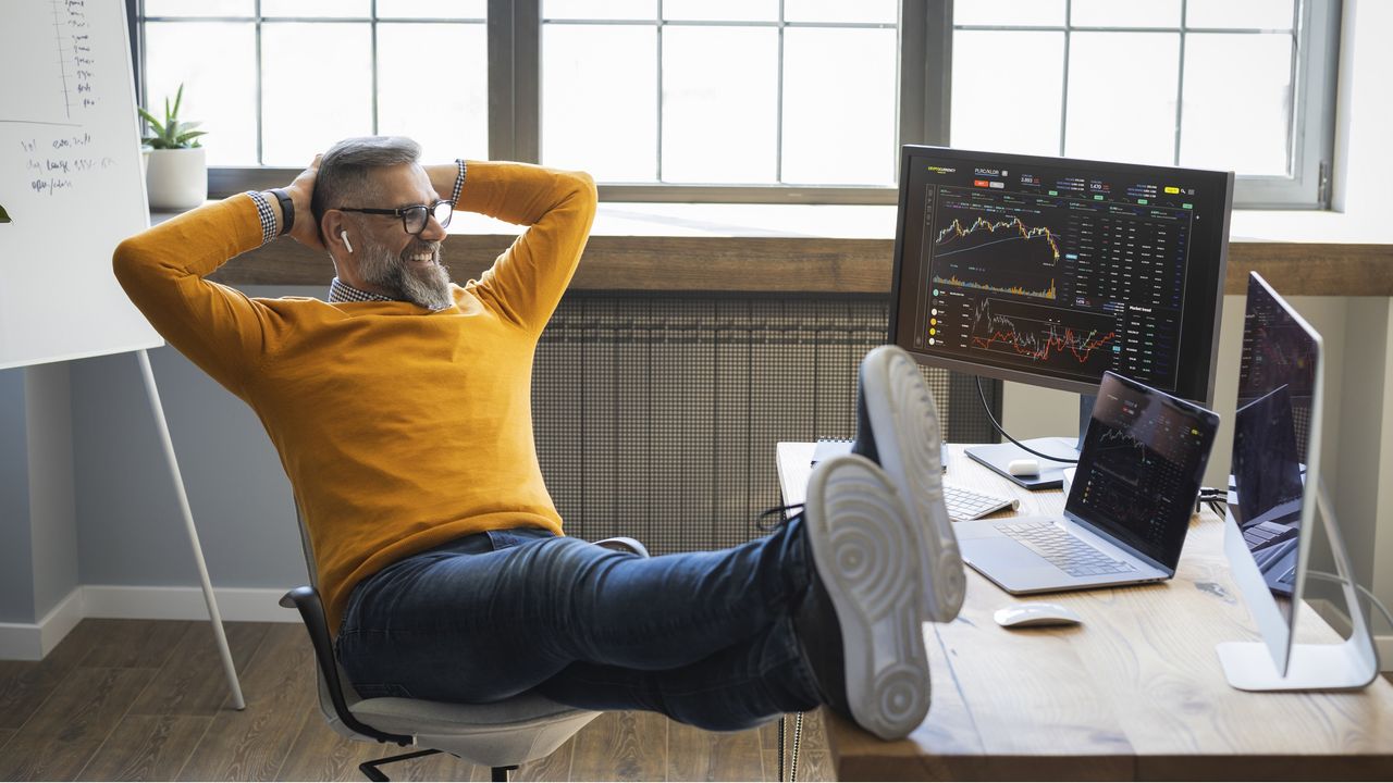 An investor looks relaxed and happy as he watches trading on his laptop with his feet up on the desk.