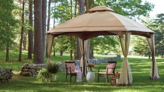 Light brown gazebo with 2 garden chairs and a grey vase with an orange flower underneath it. The gazebo is in a big garden with a stone wall and tall trees