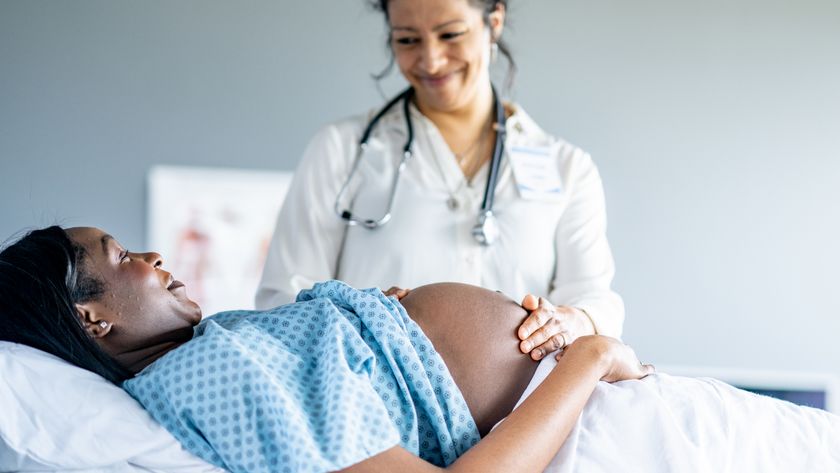 An expectant mother lays down on an exam table in a hospital gown during a routine check-up. She has her belly exposed as the doctor palpates her abdomen to verify the position of the baby.