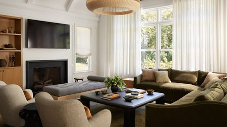 White living room with olive green velvet corner sofa, two beige armchairs, black square coffee table, grey ottoman and black fireplace