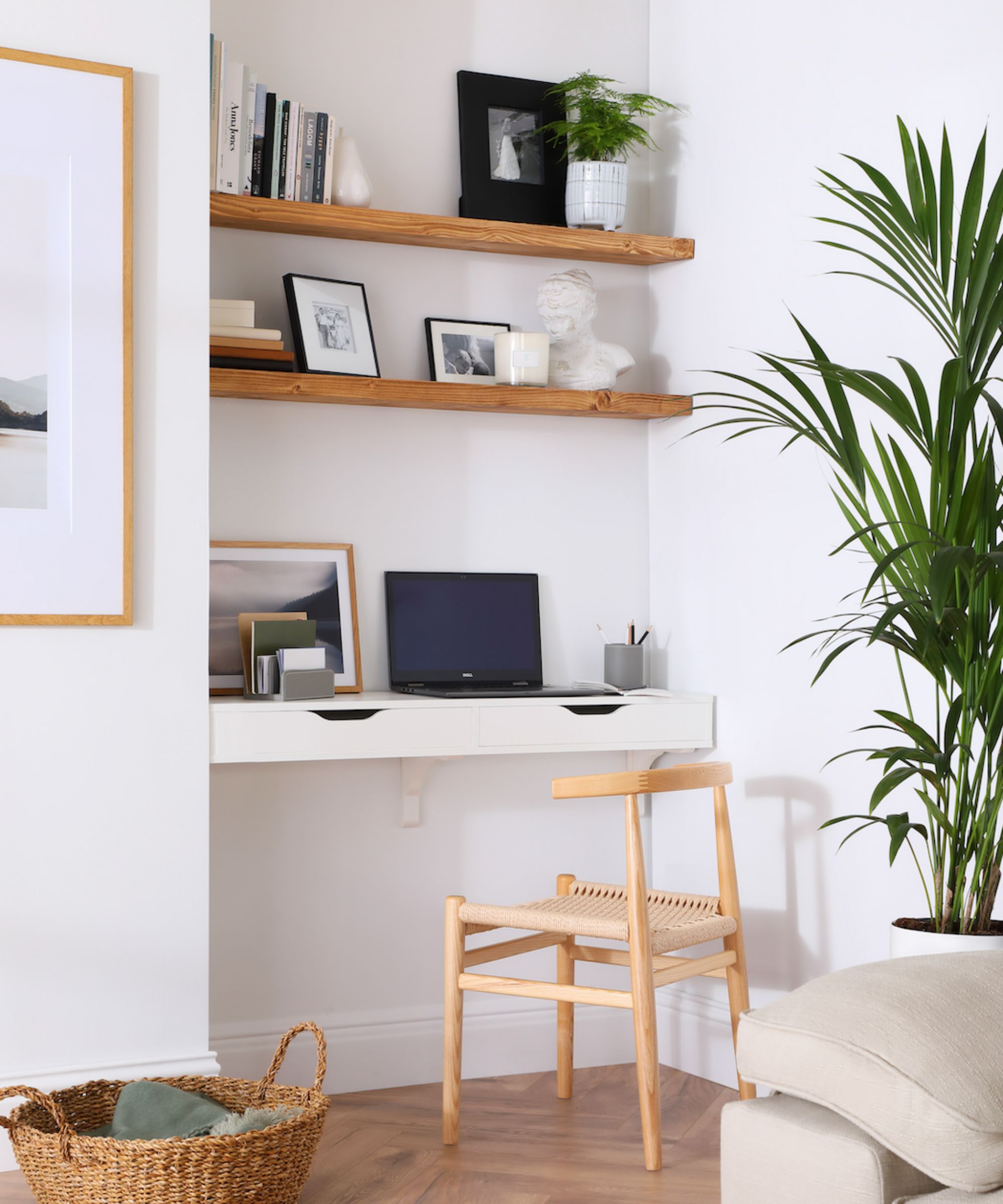 Minimalist home office nook with a white floating desk, natural wood chair, open shelving, leafy plant, and warm wood flooring beside a neutral sofa.