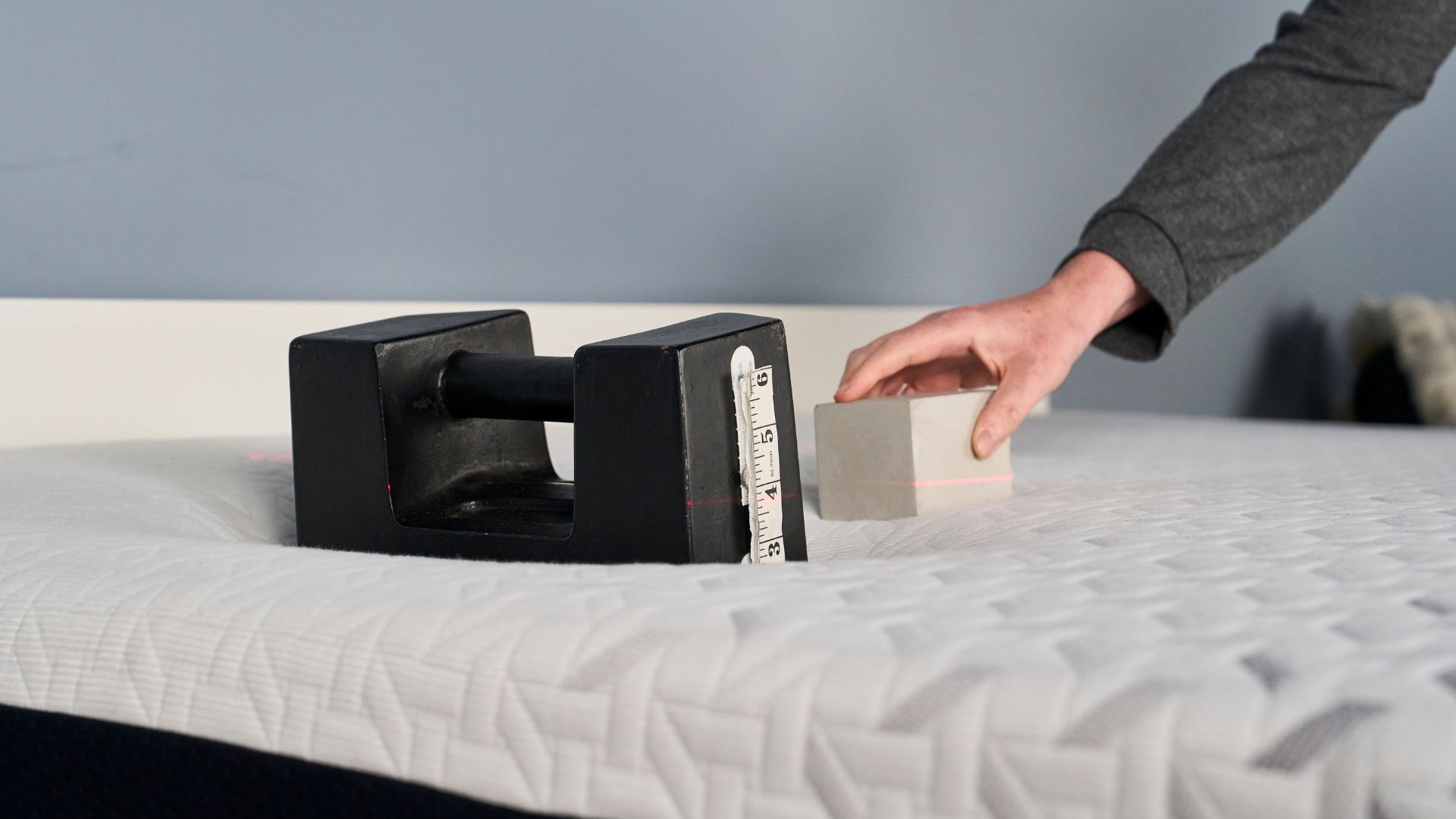 A woman'd hand places a white block to the right-hand side of a 25kg that's sinking into the shoulder region of the Nectar Premier Memory Foam Mattress. A level red laser line is projected onto both the weight and the block to measure the pressure relief of the mattress. 