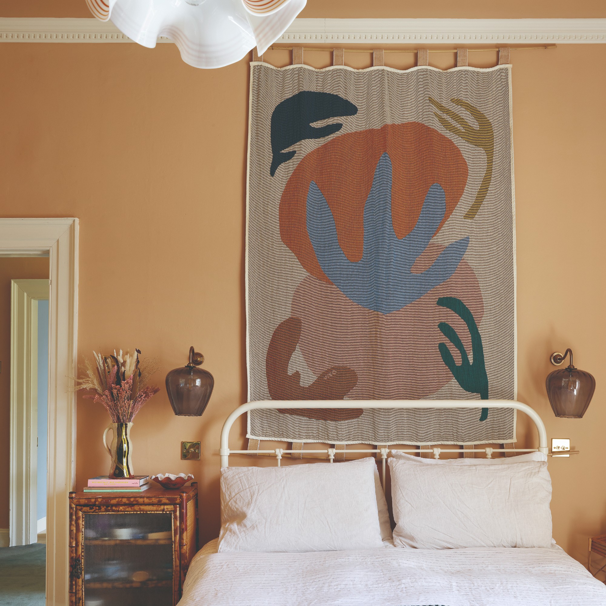 A bedroom painted in an earthy terracotta shade with an abstract wall tapestry and two wall lights on either side of the bed