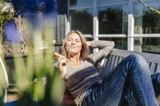 A woman in her late 40s or early 50s relaxing on a garden bench, eating a carrot.