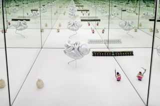 A mirrored square space with white floor and randomly placed items. In the middle of the room is an unborn baby (embossed in silver) with its umbilical cord attacked the the floor. A miniature figure in pink with a black hat holding a matchete. A miniature rabbit like figure in cream