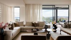 A living room with bright windows, sheer curtains, a sectional sofa with pillows and a set of circular coffee tables with vases of flowers and books around