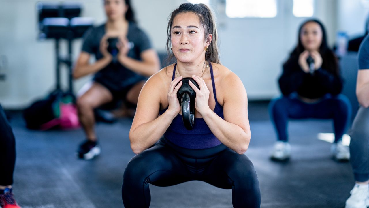 A woman performs goblet squats in a gym during an exercise class. She wears a vest and leggings and is squatting down low, with her knees wide and a kettlebell clutched to her chest. Behind her we see two other women squatting and an exercise bike in the background.