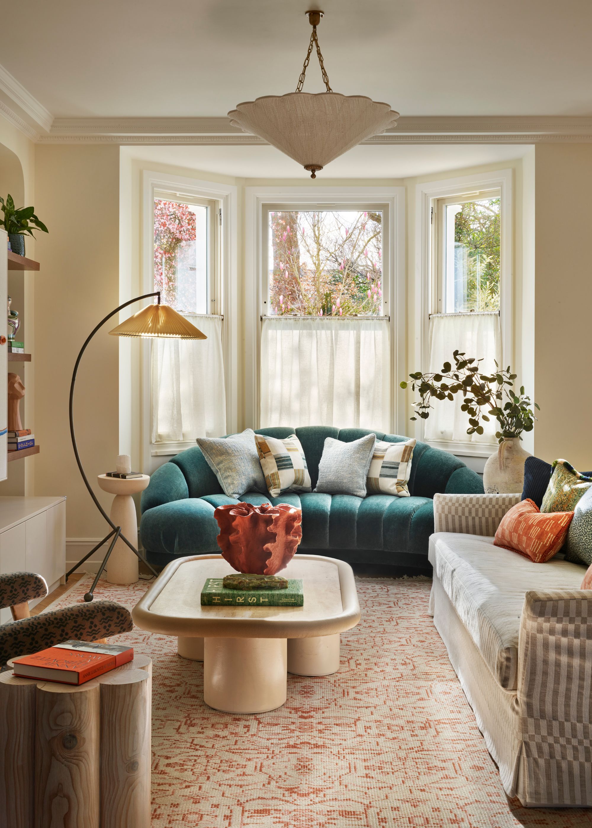 A cozy living room with warm white walls and a bay window with cafe curtains, a teal low-slung sofa in front of the window, a neutral striped sofa with a sculptural coffee table, and a patterned rug