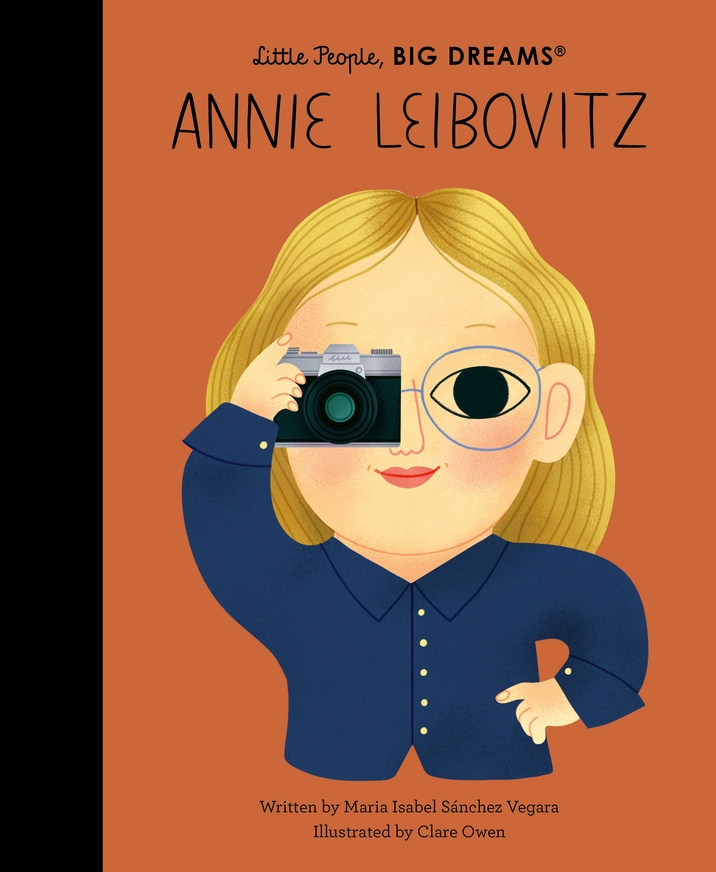 An illustration of the book cover for Little People, BIG DREAMS: Annie Leibovitz, featuring a blonde woman with glasses holding a camera up to her eye against an orange background
