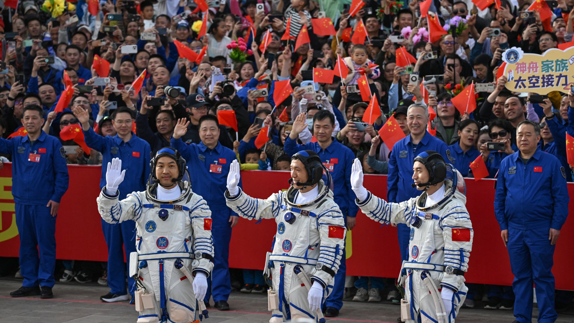 three astronauts in white spacesuits wave to a smiling crowd holding red flags