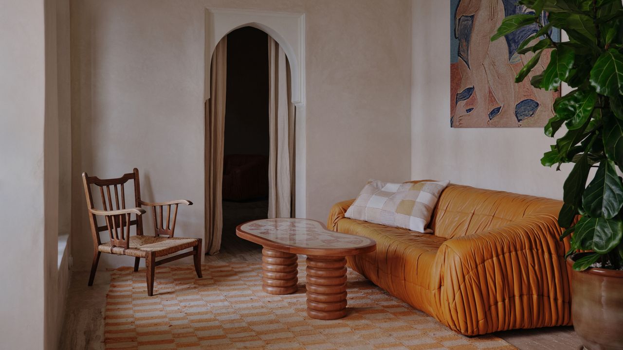 Image of a pale pink, almost beige living room. There is an orange leather sofa with a wood coffee table in front of it. The rug is a peach and off-white checkered pattern, and there is a wooden accent chair in the corner by the door frame. 