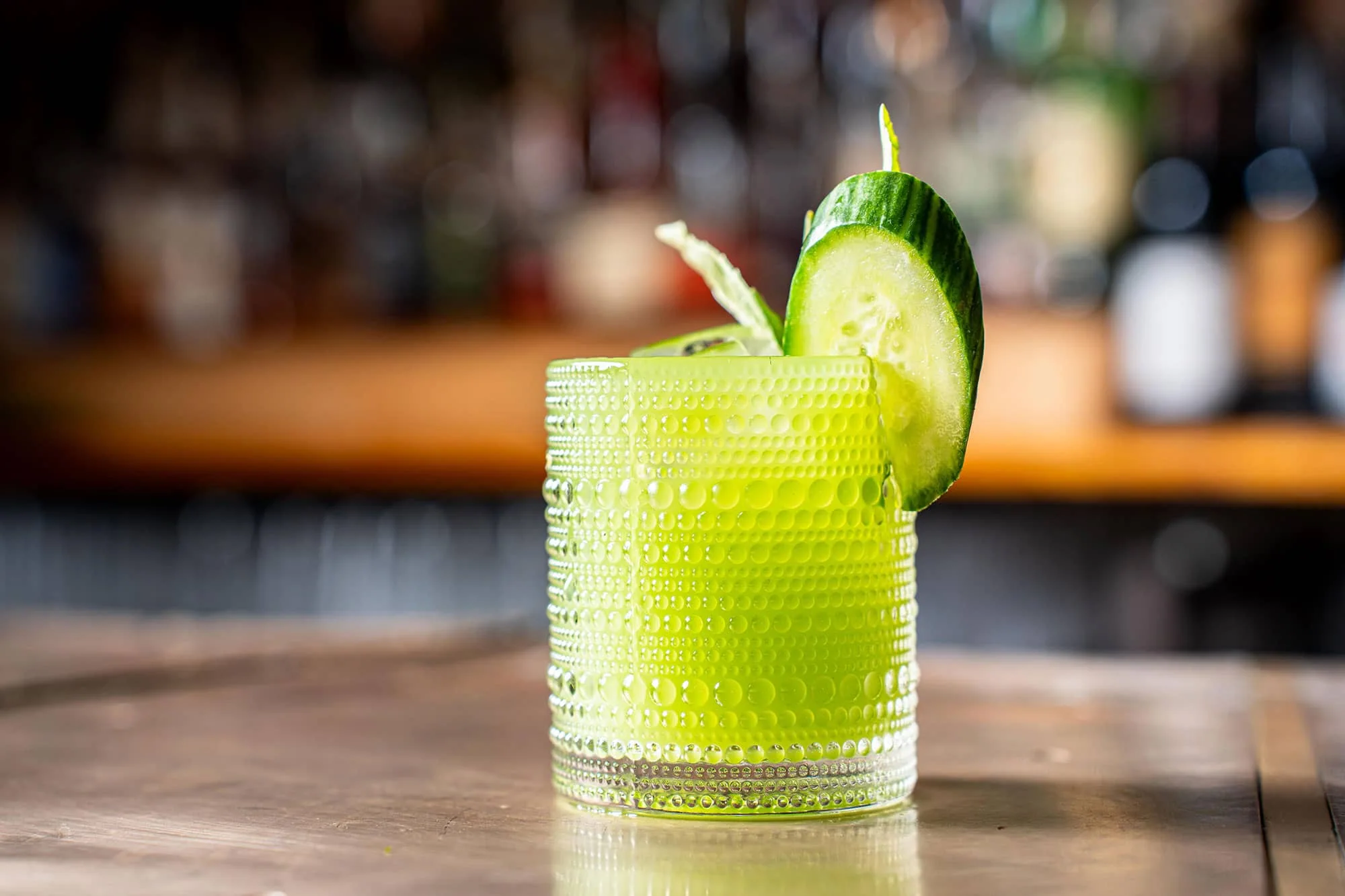 side shot of a ribbed rocks glass with green drink and a plank of cucumber as a garnish